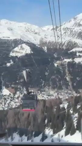 The video shows a gray and red ski gondola ascending on a set of cables. Below the gondola, a forested mountainside slopes down to a town covered with snow. In the distance, larger snow-covered mountains rise. The sky is mostly clear.  The view is from another gondola on the same cable system. There are sounds of people speaking.