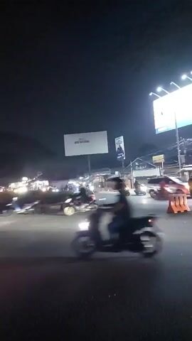 The video opens on a dark night with a wide shot of a street intersection. Several motorcycles and a red car are visible, along with traffic cones and streetlights. The camera pans right, focusing on a large illuminated sign that reads "SUVARNA SUTERA TANGERANG NEW CITY." The sign is positioned at the entrance to what appears to be a planned community or development. The street is paved, and trees are visible in the background, illuminated by the lights of the sign and nearby buildings. The overall atmosphere is that of a busy urban area at night. The video shows a typical street scene with vehicles and signage.
