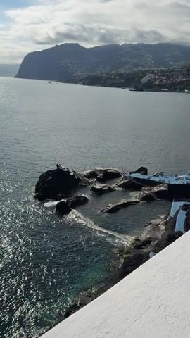 The video shows a high-angle view of a coastal landscape, featuring a rocky promontory extending into the sea. On the promontory, there is a complex of terraced pools, connected by steps and walkways. Some individuals are visible in and around the pools, suggesting they are used for swimming or leisure. The sea is calm, with gentle waves lapping against the rocks. In the background, a mountainous coastline stretches into the distance, dotted with buildings and vegetation. The sky is partly cloudy, with diffused sunlight creating a somewhat hazy atmosphere. A portion of a white structure, possibly a balcony or wall, is visible in the lower right corner of the frame, suggesting the video was taken from a building or elevated position.