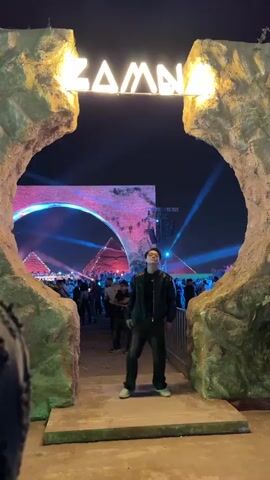 A man in a black jacket, dark pants, and light-colored shoes stands in front of a stone-like archway with the name "ZAMNA" lit up above it. He then walks through the archway, into a crowded outdoor festival. There are many people, and the background features illuminated pyramids, spotlights, and vibrant colored lighting. The man looks around and appears to dance slightly.