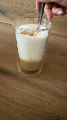 A hand with light pink fingernail polish is stirring a layered drink in a clear glass mug with a double wall. The drink has a bottom layer of caramel-colored liquid, topped with a thick layer of white foam. The spoon is silver and is actively mixing the layers together, creating ripples in the foam. The mug sits on a wooden surface, likely a table, with a visible wood grain pattern. The background is slightly blurred, but appears to be a wooden surface as well. The lighting is soft and natural.