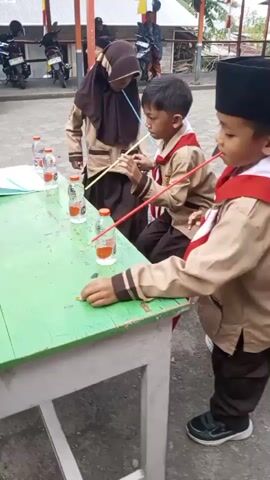 The video shows a group of children in scout uniforms playing a game outdoors. In the foreground, a table with several plastic bottles is visible. A boy in a scout uniform is reaching towards the table. In the background, other children in scout uniforms are running and participating in a game involving cones. There are also motorcycles and a building with Indonesian flags in the background. The ground is paved with interlocking bricks. The children appear to be engaged in a relay race or a similar activity.