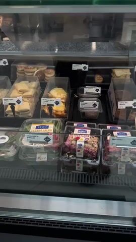 This media file shows a refrigerated display case filled with pre-packaged food items, likely in a grocery store or deli. The items include various salads, sandwiches, and other prepared meals, presented in clear plastic containers. Some of the food items are also displayed in larger trays with serving utensils. The overall scene is well-lit, suggesting an indoor setting, likely during daylight hours given the reflections on the glass.

The visible text on the labels is in Latvian. Some of the labels are in a small font, making them difficult to read entirely, but some prices are visible, such as "3.55 €", "1.55 €", "5.20 €", and "4.50 €". There are also labels indicating "JAUNUMS" (New) and a "SEASONAL" graphic with "SEZONAS SAKARS" which translates to "Seasonal Connection" or "Seasonal Offer." One label reads "CEZARA SALĀTI" which means "Caesar Salad."

The display case appears to be part of a self-service section where customers can select pre-packaged meals. The variety of food suggests a place that caters to quick lunches or grab-and-go meals. There are no people prominently featured in the frame, but the reflections in the glass suggest that this is a public space. The organization of the display suggests a focus on fresh, prepared food items available for purchase. The presence of specific food items like salads and sandwiches points towards a food service environment.