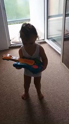 A young child stands holding a toy guitar. The child is wearing a white tank top and no pants. The child has blonde hair pulled back in a ponytail. The child is standing on a carpeted floor. Behind the child is a doorway leading outside. The toy guitar is blue and orange with colorful buttons. The guitar lights up. The child is looking at the camera and smiling. The child is holding the guitar with both hands. The child's feet are bare. The lighting is dim.