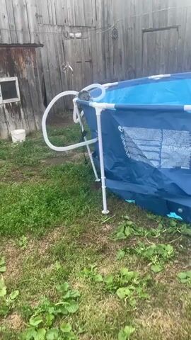 The video shows a partially assembled, blue, above-ground swimming pool in a backyard. The pool has a white metal frame and a blue liner. A hose is attached to the pool. The backyard has a wooden fence and some overgrown grass and weeds. There is a ladder leaning against the fence in the background.