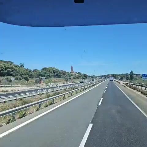 The video, shot from a moving vehicle, shows a multi-lane highway under a bright, clear blue sky. On the opposite side of the highway, a hill dotted with trees and low vegetation rises. Perched atop the hill is a village or town dominated by a tall, cylindrical stone tower with a red-tiled roof. A black van travels on the highway opposite the camera. The road surface appears to be in good condition, with clear lane markings. The overall impression is a sunny, clear day with a picturesque village visible from the highway.
