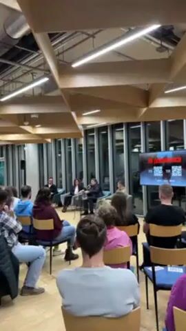 The video shows an audience seated and listening to a panel of four people during what appears to be a discussion or presentation. The panel members are seated on chairs at the front of the room. The audience is seated in rows of chairs facing them. The audience includes people of different ages and genders, with varied clothing.  A screen behind the panel displays text and QR codes. The room has a modern design, featuring a high ceiling with wooden beams, a large window, and artificial lighting. The overall setting suggests an educational or community event.