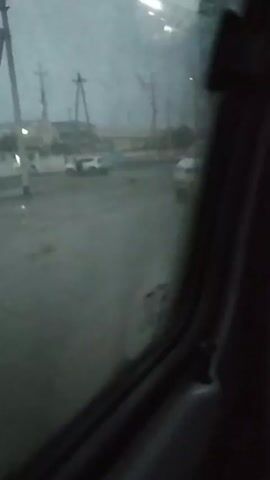The video shows a view from inside a vehicle, looking out a side window. The glass is somewhat fogged or wet. The outside scene is blurry but appears to be a gray, overcast day. There are utility poles with wires running along them. A couple of buildings are visible in the distance, along with a few lights. The road outside looks wet. The camera does not move much.