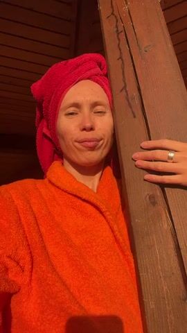 A woman with a red towel wrapped around her head and an orange bathrobe stands outside. She blinks and smiles at the camera. A wooden beam is to her right, and a hand with a ring is resting on it. The background shows a brick wall and some stacked wood. The lighting suggests it is daytime, possibly late afternoon, with sunlight casting shadows.