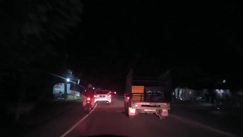 The video depicts a nighttime scene from the perspective of a driver following other vehicles on a paved road. A motorcycle and a white car are positioned ahead, with a small truck directly in front, its rear illuminated by red taillights. The truck's cargo includes containers or boxes. The road is lined with buildings, with signs visible on some, including one for "HENDRI CELL." Illumination comes from vehicle lights and sparse streetlights.