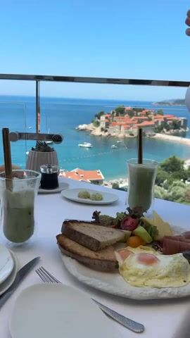 This media file showcases a picturesque breakfast setting with a stunning coastal view. In the foreground, a white tablecloth is laden with a delicious-looking breakfast spread. This includes two glasses of iced green beverage with straws, a plate of toasted bread slices, a salad with cherry tomatoes and cheese, a serving of cured meat, and two fried eggs. Accompanying the meal are a bowl of assorted bread rolls, a small plate with two portions of butter, and a small container of what appears to be jam or sauce. Silverware and white plates are also arranged on the table.

The background features a breathtaking panoramic view of Sveti Stefan, a historic islet in Montenegro, connected to the mainland by a narrow isthmus. The turquoise waters of the Adriatic Sea surround the island, dotted with a few sailboats. The islet itself is adorned with terracotta-roofed buildings, creating a charming and idyllic scene. Beyond the sea, lush green hills and more coastal architecture are visible, under a clear blue sky. The overall atmosphere is one of serene luxury and enjoyment, suggesting a leisurely morning meal in a beautiful location. The time of day appears to be morning or early afternoon, with bright sunlight indicating pleasant weather. No people are visible in the frame, emphasizing the peaceful and tranquil setting. There is no visible text in the media file.