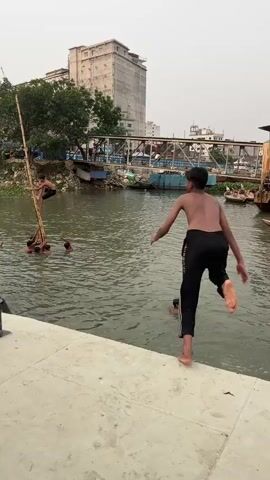 The video shows a group of young boys playing and swimming in a river or canal in an urban setting. One boy is climbing a tall, slender bamboo pole that is being held upright in the water by several other boys who are submerged up to their chests. Another boy jumps into the water from a concrete embankment. The backdrop includes buildings, a bridge, and some boats docked along the canal. A man in a white outfit is visible near the edge of the embankment, and there is some vegetation along the water's edge. The sky is overcast, suggesting it might be early morning or late afternoon.