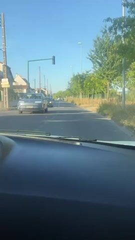 The video shows a view from inside a vehicle looking out the windshield. The vehicle is on a paved road with a clear blue sky above. On the left side of the road, there are houses and telephone poles. On the right side of the road, there is a sidewalk with trees and a fence. Several cars are visible on the road, some moving and some parked. There is a traffic light ahead and a pedestrian crossing. The sun appears to be shining, casting shadows. The overall environment appears to be a residential area.