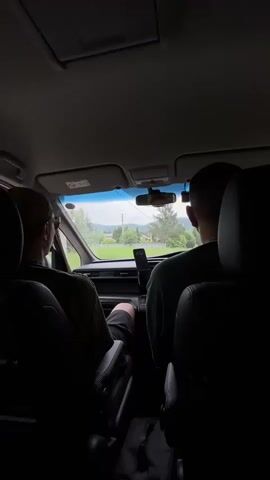 The video shows the interior of a car from a rear perspective. Two individuals are seated in the front, with their backs to the camera. The person in the driver's seat, on the left, is wearing a green t-shirt and shorts, and has sunglasses and a cap on. The passenger, on the right, is wearing a green t-shirt. The car appears to be moving on a road through a rural or suburban area, with greenery and some buildings visible outside the windshield. A smartphone is mounted on the dashboard.