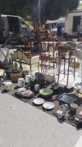 The video pans across an outdoor flea market. In the foreground, a dark brown tarp is laid out on the ground, displaying various items for sale. These include several plates with red accents, a green bowl with a woven pattern, a metal grinder, a metal tray with a scenic image, and various decorative objects such as masks and gold-colored figurines. Behind these, a collection of white cylindrical containers and a small, gray, rectangular item are visible. Further back, an ornate, upholstered armchair and matching chair are displayed. Behind these chairs, a tall, multi-tiered metal shelf unit stands, with a few items placed on its shelves. Two tall, wooden stools with cushioned seats are placed in front of this shelf. Several white, translucent stools are arranged to the left of the shelf. To the right of the shelf, a modern, chrome-plated bar stool with a white seat is visible. In the background, several white tents and vans are set up, indicating the presence of more vendors. People can be seen browsing and interacting in the background, with some appearing to be vendors tending to their stalls. The overall environment suggests a sunny day at a bustling outdoor market.