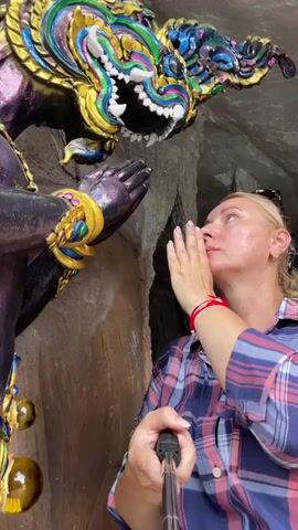 A blonde woman with a red bracelet on her left wrist holds a selfie stick and points it towards herself and a statue. She wears a plaid shirt and has her right hand near her mouth, as if whispering. The statue appears to be a mythical creature with a colorful, ornate head and a purple body. The background consists of a rocky cave-like environment.