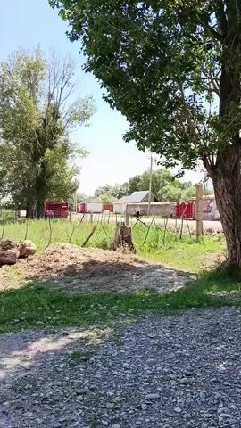 The video shows a rural scene on a sunny day. The foreground is a gravel path, with a pile of dirt and straw to the left. Behind this, a wooden fence runs across the middle ground, with grassy fields and trees on either side. In the background, there are several buildings, including houses and a red structure that appears to be a truck or trailer. The sky is clear and blue. The camera pans slowly to the right, offering a wider view of the landscape.