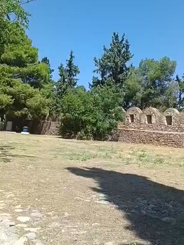 The video shows a long stone wall with arched openings. The wall is made of rough, irregularly shaped stones and appears to be quite old. The openings in the wall are evenly spaced and have rounded tops. Behind the wall, there are trees and a clear blue sky. In the foreground, there is dry, patchy grass and a long shadow cast by an unseen object. The camera pans slowly across the scene, providing a view of the wall from different angles. There are no people visible in the video.