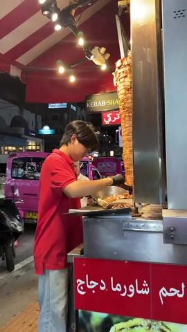 A person wearing a red shirt and black gloves is preparing food at a shawarma stand. They are using a knife to slice meat from a vertical rotisserie. The stand has red and white striped awning and various signs with text in Arabic and English. In the background, there are other food stalls, a pink vehicle, and people walking. The scene appears to be at night, with streetlights and illuminated signs.