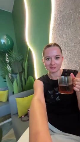 A woman with blonde hair pulled back in a bun, wearing a black t-shirt and light-colored pants, holds a clear glass mug of dark liquid to her lips and drinks from it. She is seated on a gray sofa with bright yellow throw pillows. Behind her, a large green balloon is tied to a leafy plant. The room is decorated in shades of green with a wavy yellow and pink neon light fixture on the wall. To the left of the sofa, a small green side table is visible. The woman is positioned in the foreground, and the video appears to be a selfie taken by her.
