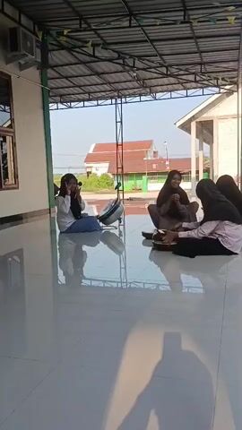 A video captures four young women sitting on the floor beneath a covered outdoor area. The floor is highly reflective, showing the reflections of the women, a fan, and the surrounding environment. Three of the women are wearing hijabs. One woman on the far left, wearing a white top and blue jeans, is looking towards the camera and making a hand gesture. In the center, two women are seated facing each other. The woman on the left in this pair is wearing a brown top and appears to be holding something brown and round in her hands. The woman on the right is wearing a dark hijab and a light pink top. To the right of these two, another woman with a black hijab and a black top is seated, her back mostly to the camera. In the background, there are buildings and a clear sky, suggesting daytime. A white fan is positioned on the floor to the left of the group.
