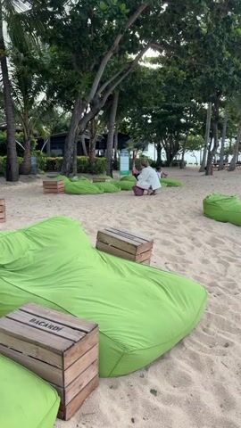 A drone shot pans across a beach with green beanbag chairs and wooden crates on the sand. A few people are visible in the background near trees and lounge chairs. The water is visible in the distance with a cloudy sky overhead.
