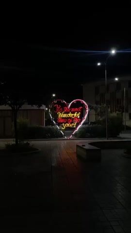 A large heart-shaped neon sign is illuminated in red and pink lights, displaying the text "It's the most wonderful time of the year!" The sign is situated in a public outdoor space at night. The ground is paved with interlocking bricks. In the background, there are buildings and streetlights. To the left of the heart sign, there is a small tree and some shrubbery. To the right, there is a bench. The scene is dark, with the neon sign providing the primary illumination.