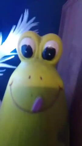 This is a close-up video of a yellow toy frog with large eyes and its tongue sticking out. The frog is illuminated by a string of bright blue LED lights that appear to be attached to a plant or some sort of decorative display. The background is dark and out of focus, with some hints of green and white. The video is shaky and the lighting is inconsistent, making it difficult to discern fine details.