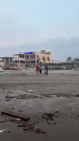 The video shows a beach with a hotel in the background. The beach is cluttered with debris, including driftwood and trash. There are several small fishing boats scattered along the shore, along with small huts elevated on stilts. Two people are walking on the beach, and a third person is riding a bicycle. The sky is overcast and gray.
