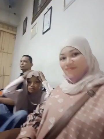 This is a video still of a family posing for a photo. Three people are visible: a man in the background, a young girl in the middle, and a woman in the foreground. The man is wearing a black polo shirt and jeans. The girl is wearing a light pink shirt and a beige hijab. The woman is wearing a light pink hijab and a patterned dress. The family is sitting on a couch in what appears to be a living room. The walls are white, and there are framed pictures on the wall. The lighting suggests it might be evening or indoors. The mood appears to be relaxed and happy, with the woman and girl smiling at the camera. There is no visible text in the image. The overall scene depicts a casual family moment.
