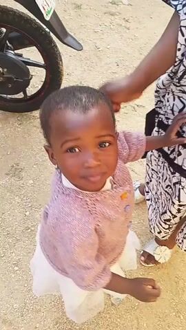 A young Black girl, approximately two years old, is seen standing in a dirt area next to a woman. The girl is wearing a white dress, pink cardigan with colorful buttons, and earrings. The woman is wearing patterned pants and sandals. A motorcycle is parked behind the girl and the woman. A hand brushes the girl's head with what appears to be a hairbrush. The girl is looking at the camera. The girl raises her hand to her head.