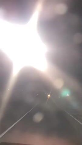 The video shows a nighttime scene, possibly from inside a vehicle, with a bright light source dominating the upper left portion of the frame. The light is a strong, starburst-shaped glare, suggesting a streetlamp or headlights. There are visible bokeh effects, indicating the presence of water droplets or dust on a lens. In the distance, two small, yellowish lights are visible, possibly other streetlights or vehicle lights. The background is dark and indistinct, with faint lines suggesting a road or pathway. The overall impression is of motion and low visibility.