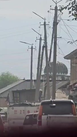 The video begins with a close-up of power lines and poles, then pans out to show a street scene. Several cars are on the road, some parked and some in motion. There are also buildings on either side of the street, and trees with green leaves. A mosque with a green dome is visible in the background. The scene appears to be a residential or commercial area with moderate traffic. The overall atmosphere is bright and sunny.