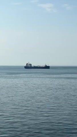The video shows a vast, calm body of water with a clear blue sky above. A large cargo ship, appearing to be a tanker, is the primary subject, positioned centrally in the frame and sailing horizontally. The ship has a long, slender hull with a white superstructure at the front and a smaller structure towards the stern. The water is a deep blue-grey with gentle ripples, suggesting a serene and tranquil atmosphere. The sky is a pale blue, with a few wispy clouds scattered near the horizon, indicating fair weather. The scene appears to be captured during daylight, likely morning or afternoon, given the bright illumination.

In the latter part of the video, the perspective shifts slightly, revealing a white railing in the foreground. A circular green sign with a white symbol resembling a lifebuoy is attached to the railing. This suggests that the video is being filmed from another vessel, possibly a ferry or a smaller boat, moving in the same general direction as the cargo ship or observing it from a parallel path. The overall impression is one of peaceful travel on the open sea, with a sense of scale emphasized by the solitary cargo ship against the expansive seascape. There is no discernible text in the video.