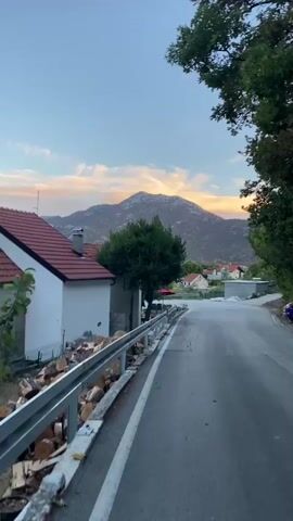 The video shows a paved road with a white line on the left side, curving to the right. To the left of the road, there is a white building with a red-tiled roof, a metal guardrail, and a pile of cut logs. A large green tree stands behind the guardrail. In the background, several houses with red-tiled roofs are visible, along with a large mountain. The sky is blue with streaks of orange and yellow clouds, suggesting either sunrise or sunset. The scene appears to be a rural or semi-rural area.