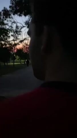 The video shows a first-person perspective of someone riding a scooter or motorcycle. The rider is wearing a red garment, and a portion of the vehicle's red body and a mirror are visible in the foreground. The scene is set at dusk, with a twilight sky displaying hues of orange and blue. The road ahead is dark, with trees lining both sides. In the distance, on the left side of the road, a lone figure is walking away from the camera. There are streetlights visible along the road. The overall atmosphere is peaceful and serene, with the natural beauty of the sunset as a backdrop.