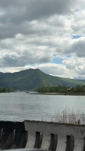 The video shows a wide river bordered by green mountains and snow-capped peaks in the background. The sky is partly cloudy. The camera is positioned behind a concrete barrier, suggesting it was taken from a bridge or elevated location. A few power lines are visible overhead. There are some signs of civilization on the shore opposite the camera, including a few buildings. The water is rippling, indicating a slight breeze. The video appears to have been taken from a moving vehicle, possibly on a road running alongside or across the river.