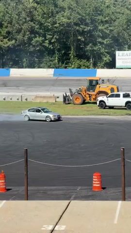 A silver sedan is seen drifting on a paved track, emitting smoke from its tires. In the background, a yellow loader and a white pickup truck are parked on the grass. There are several tents set up and people are gathered around them. A large banner displaying advertisements hangs above the track. Orange traffic cones and red barriers are visible on the track, marking the course. The scene appears to be an outdoor event or competition.
