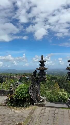 The video shows an ornate stone gateway overlooking a lush landscape under a blue sky with puffy white clouds. The gateway features intricate carvings and tiered towers on top of each post. The paved area in front of the gateway has a patterned design. Beyond the gateway, the view opens up to a green expanse with trees, houses, and distant hills. The sky is bright, and the overall impression is one of serene beauty and cultural richness.