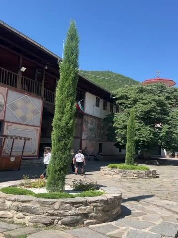 The video shows a courtyard with stone paving and several buildings. In the foreground, there are two tall, slender cypress trees in stone planters. In the middle ground, there are people walking and standing. One person in the center is wearing a white shirt and black shorts, and two others are wearing black. There is a large tree with green leaves to the right, and a building with a red tiled roof behind it. The buildings have white stone walls and some have colorful murals. The sky is blue and clear, with some trees and mountains visible in the background. The video pans slowly across the courtyard, showing different views of the buildings and the people.