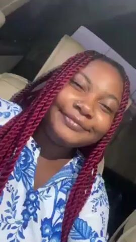 A young woman with reddish-purple braided hair is in a car. She is wearing a white shirt with a blue floral pattern. She is looking at the camera and smiling. She has a nose ring. She is touching her hair with her right hand. There is a car seat behind her.