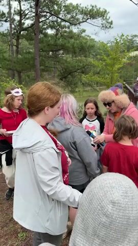 The video shows a group of children and one adult outdoors in a wooded area with a body of water in the background. The adult, wearing a pink jacket and a fur-lined hood, holds a microphone and appears to be speaking to the children, some of whom are holding papers. The children are of various ages and are dressed casually. Some are looking at the adult, while others are looking away or at each other. The ground is covered in pine needles, and there are trees and some wooden structures visible in the background.