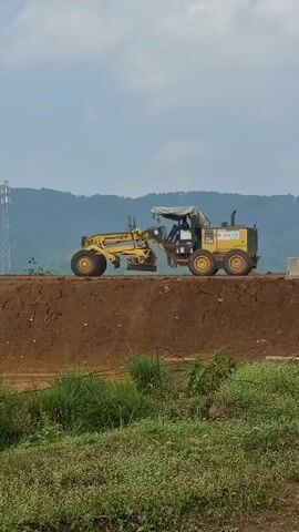 A yellow grader with a person inside is seen from the side, moving along a dirt embankment. The grader is equipped with a large blade in the center, which appears to be in contact with the ground, levelling the surface. In the background, rolling hills are visible under a cloudy sky, and a tall cell tower stands on the left. The foreground shows green vegetation. The grader is the primary focus of the image, actively engaged in its construction task.