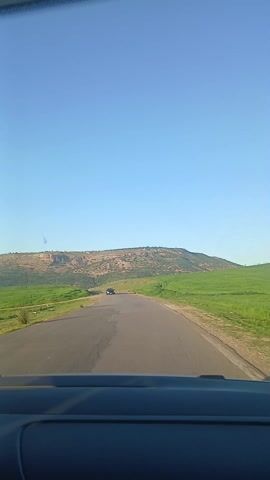 The video is filmed from the perspective of a vehicle driving on a paved, two-lane road. The bottom portion of the frame shows the top of the car's dashboard and the windshield, indicating an interior view.

The road ahead is relatively straight, with a slight uphill gradient in the distance. On both sides of the road, there are wide, lush green fields, suggesting agricultural land or pastures. In the far background, a prominent, elongated hill or plateau stretches across the horizon. This hill appears drier and rockier than the foreground fields, with patches of brown and sparse vegetation, giving it a somewhat barren appearance. The sky above is clear and a vibrant, light blue, without any clouds.

Throughout the video, other vehicles are visible on the road, traveling in the opposite direction.
- At the 0:00 mark, a light-colored vehicle is seen in the distance, slowly approaching.
- At 0:01, this vehicle passes out of frame to the left.
- Around 0:03, a dark-colored sedan appears further down the road, also heading towards the camera's position.
- By 0:05, this dark sedan has moved closer, about to pass the camera.

The overall impression is of a drive through a scenic, rural landscape on a clear, sunny day.