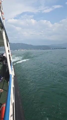 The video begins with a close-up view of the side of a white boat. People are visible on the deck of the boat, some standing and some sitting. The boat is moving through green water, creating a wake. In the distance, a hazy coastline with mountains can be seen under a cloudy sky. The camera pans to the right, revealing more of the expansive body of water and the distant shore.