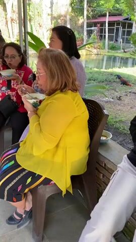 A group of people are eating from bowls in an outdoor setting. The individuals are dressed casually and appear to be enjoying a meal together. The background shows lush greenery and a body of water.