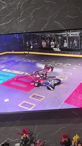 A televised robot combat event is displayed on a screen. In the arena, a large, mechanical arm with a circular saw at its end is positioned above a smaller, blue robot that appears damaged and is lying on its side. The arena is marked with colored sections and logos. In the foreground, several people are visible on a platform, seemingly operating the robots. One person in the center is holding a controller. The background of the arena shows spectators in elevated seating. Below the screen, several anime figurines are arranged. The scene is framed by a textured surface.