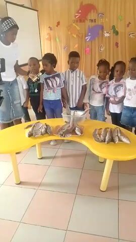 This video shows a group of young children, approximately four to six years old, standing in a line in front of a yellow table. They are all facing the camera, and most are holding up pictures of fish. On the table, there are several real fish displayed, some of which appear to be gutted. A woman, presumably a teacher, is standing to the left of the children, holding a phone and appearing to record the scene. The background is decorated with a yellow curtain featuring an "Under the Sea" theme, with cutouts of sea creatures and bubbles. The children are dressed in casual clothing, and the floor is tiled.