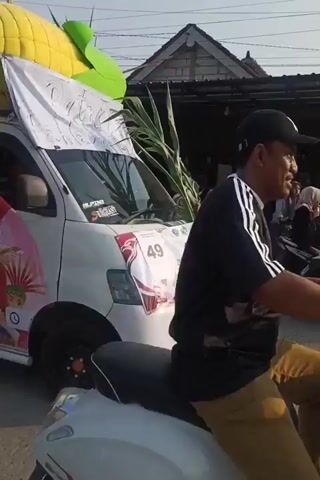 This is a video taken from a low angle looking up at the side of a white pickup truck participating in a parade. The truck is decorated with an inflatable corn on top, a large white banner with red lettering that reads "MARAH MERDEKAAN" (likely "Independence Parade" or similar), pink floral garlands, and green foliage. The sides of the truck are adorned with colorful illustrations of people in traditional attire. Several people, mostly young women wearing headscarves, are visible in the truck bed, waving to the camera and appearing to be part of the parade. The truck is moving, and other vehicles, including motorcycles, are visible in the background, suggesting an outdoor street setting. The sun is bright in the sky.