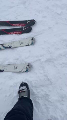 The video opens with a close-up of two pairs of skis standing in the snow and a black ski boot. The top pair of skis are black and red with the brand name "FISCHER" displayed, while the bottom pair are white with a turquoise and purple design and the brand name "ROSSIGNOL LTD." The camera then pans up to show a snowy landscape with scattered pine trees and a mountain range in the background, partially obscured by heavy fog. A coin-operated telescope stands prominently in the foreground. The camera continues to pan right, revealing a small red building with a sign. As the camera movement ends, a group of people in ski attire are seen standing on the snow, seemingly preparing to ski. They are facing away from the camera, and their interactions, if any, are not visible. The overall scene depicts a snowy mountain resort in overcast weather conditions.